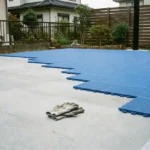 自宅の庭でコンクリート基礎の上に青いNEXCOURTのモジュラータイルをDIYで施工している様子。手前には作業用手袋が置かれている。